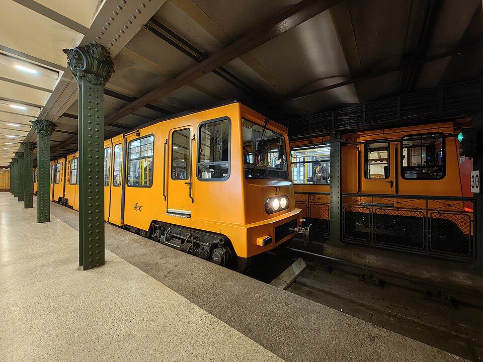 Platform of the Hosok tere station on Budapest's M1 Millennium Underground line, with a small yellow/orange train car beside decorative green cast-iron columns supporting the vaulted ceiling