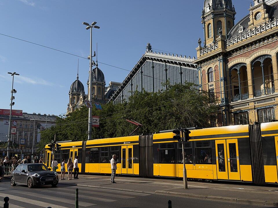 Modern yellow Budapest tram running past Nyugati railway station, with pedestrians, a car, and the station's glass-and-iron facade in the background