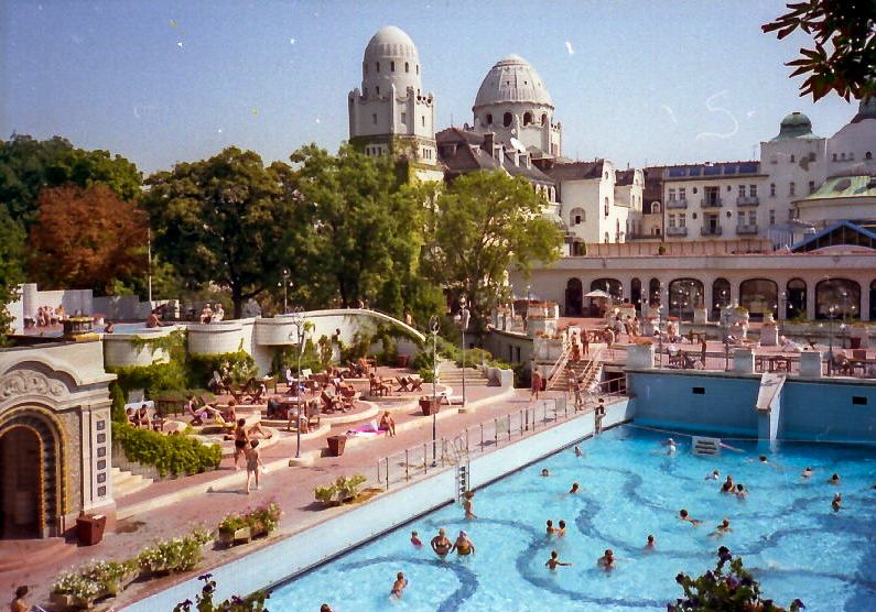 Outdoor wave pool at Gellert Baths in Budapest with bathers in the water and the Art Nouveau domed building rising behind terraced gardens