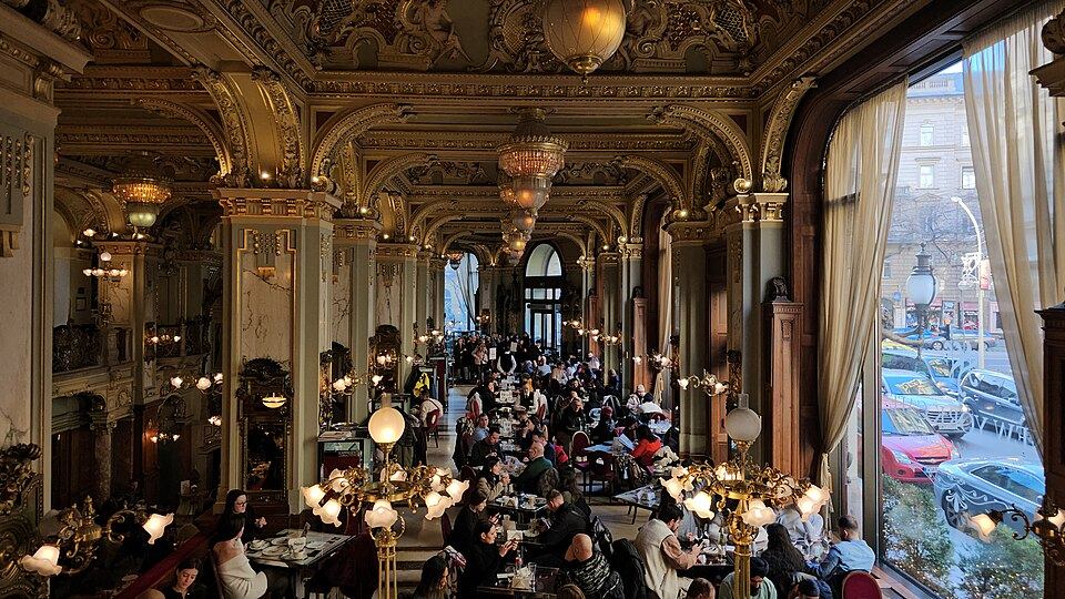 Gilded, frescoed interior of the New York Cafe in Budapest with ornate columns, chandeliers, and patrons seated at tables near tall curtained windows overlooking the street