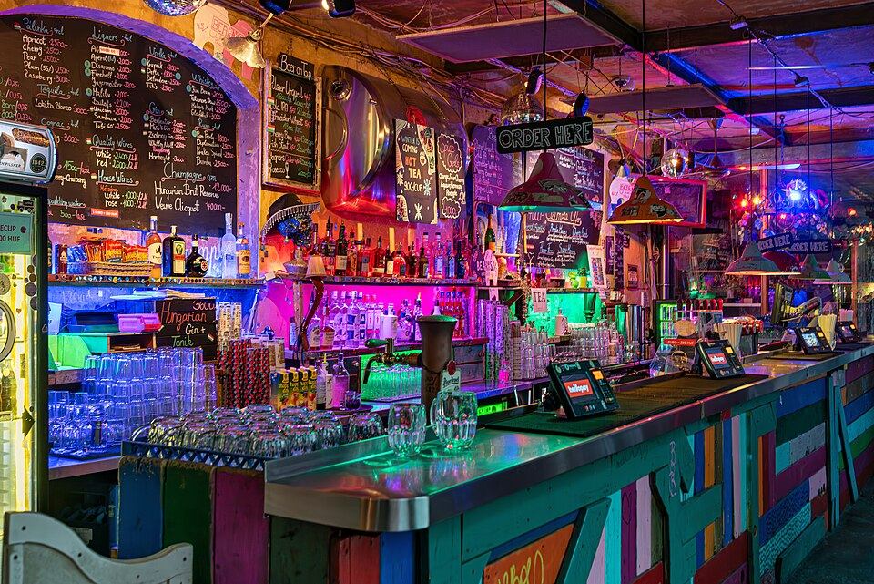Colorful, eclectically decorated bar counter inside Szimpla Kert ruin bar in Budapest, lit with neon and string lights, chalkboard drink menus on the walls